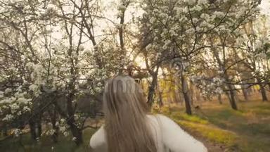 穿着毛衣、牛仔裤的迷人年轻女子在盛开的苹果树间翩翩起舞。 日落，春天的果园和